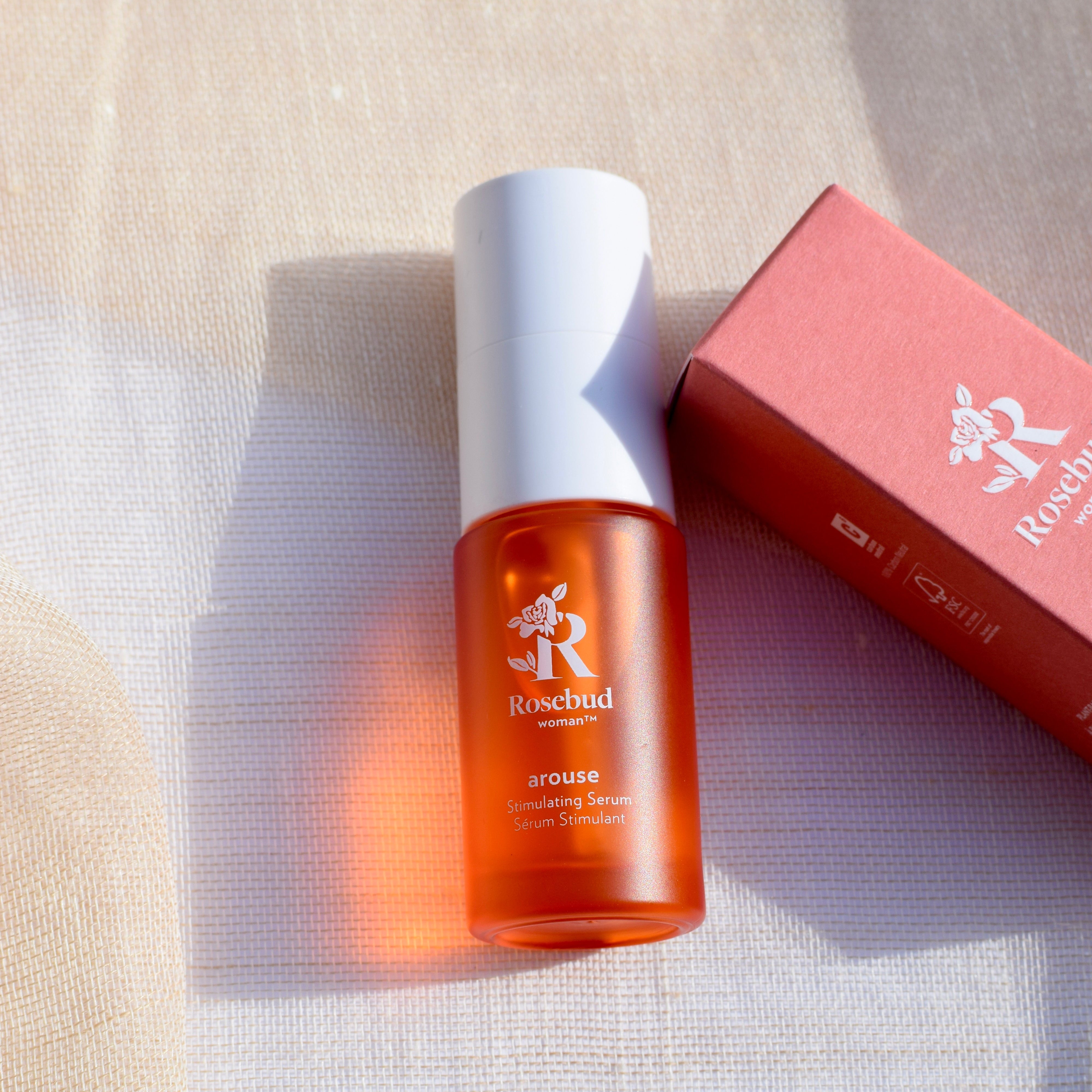 Rosebud woman arouse stimulating serum   bottle glowing in the sun with a box on a textured beige linen surface 